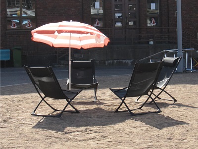 Kuva aurinkotuoleista ja aurinkovarjosta hiekalla punaisen tiilitalon edessä