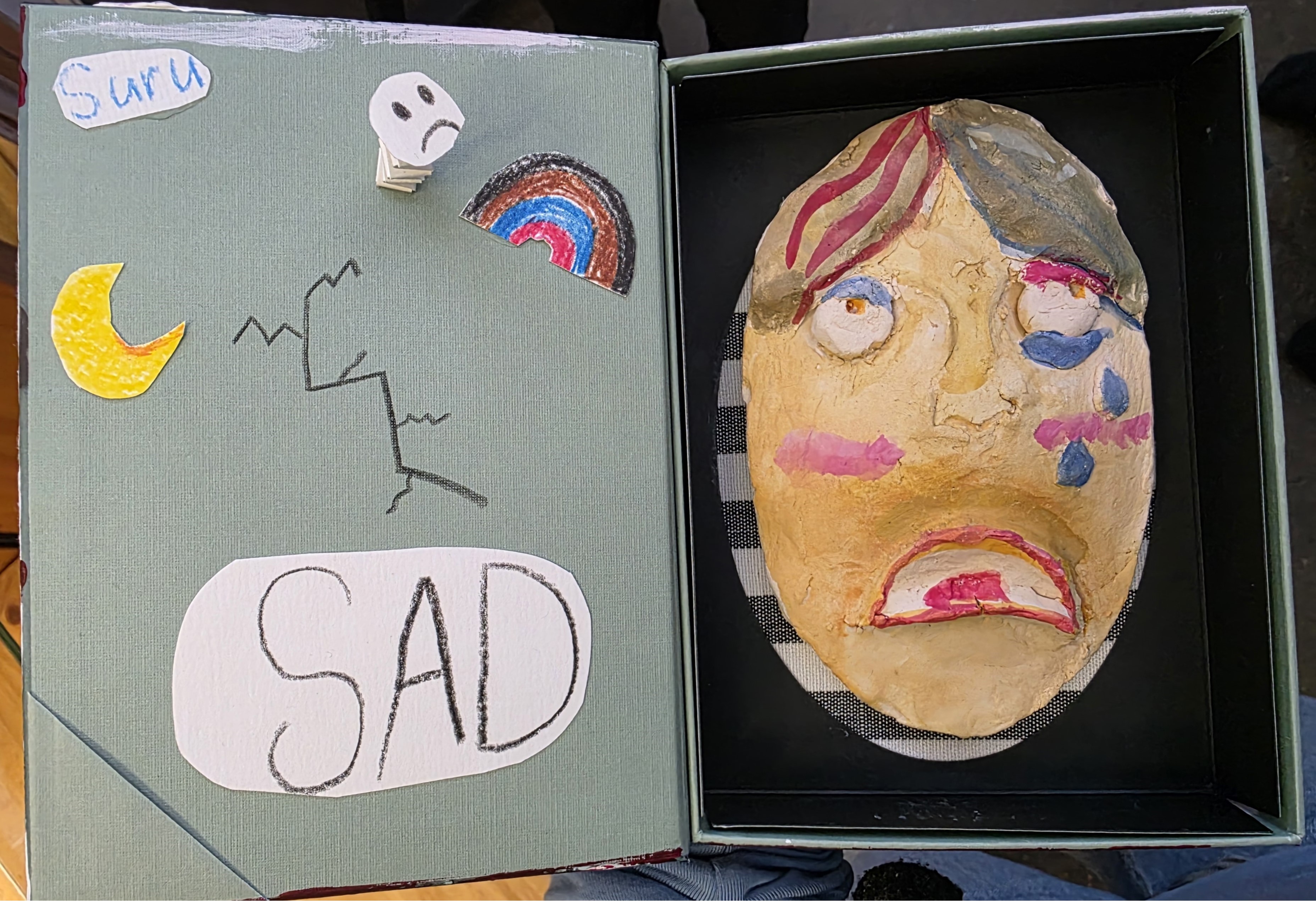 Laatikossa keramiikkakasvot, vieressä lukee suru ja sad sekä on kuvia salamasta, kuutamosta ja sateenkaaresta