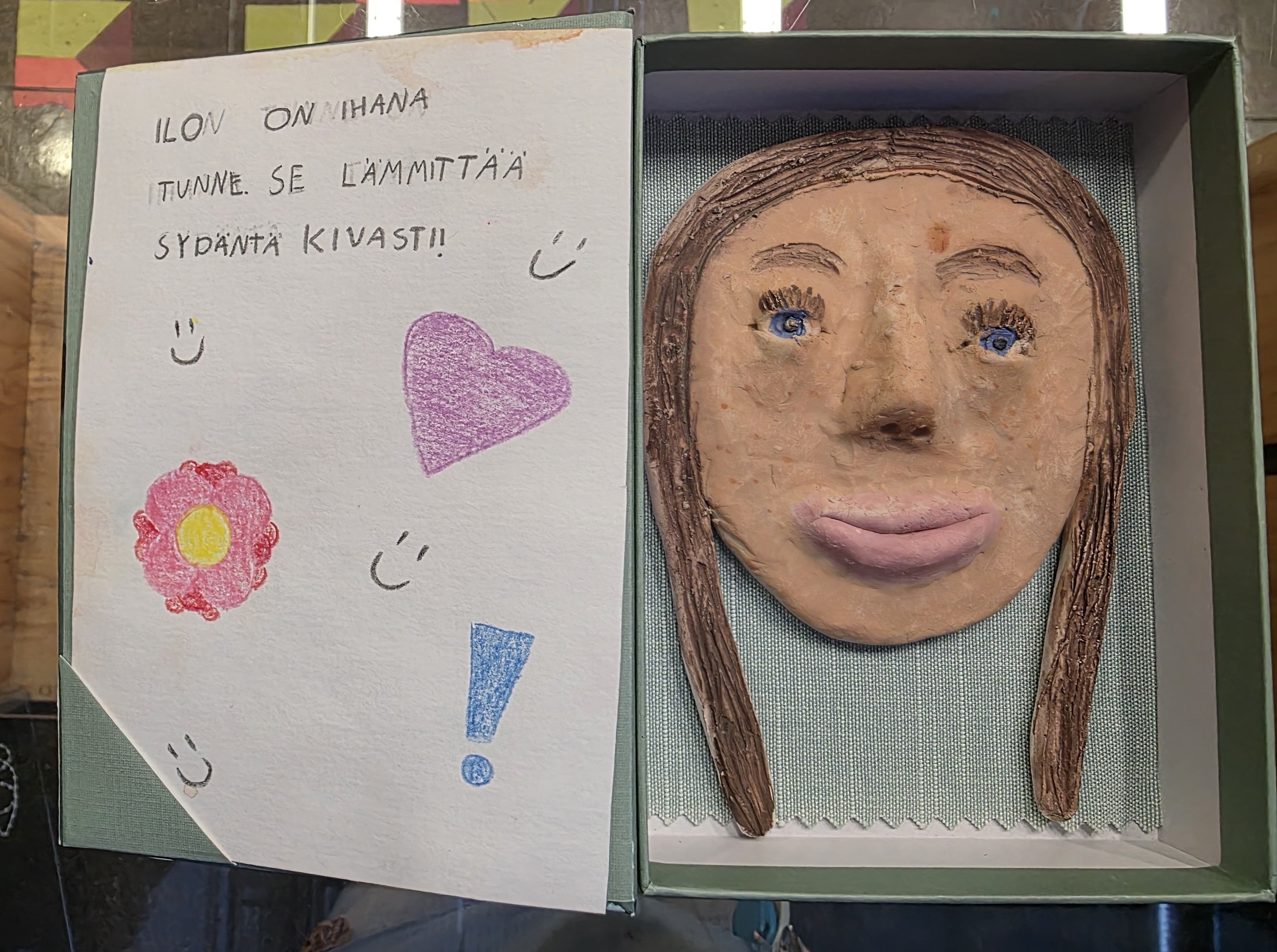 Laatikossa keramiikkakasvot, vieressä lukee: ilo on ihana tunne se lämmittää sydäntä kivasti