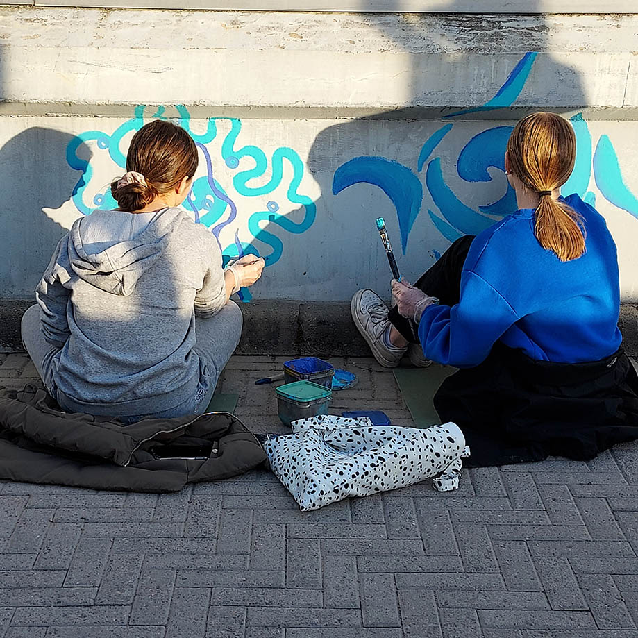 Kaksi oppilasta istuu maassa selin kameraan ja maalaalaa siveltimillä sinisiä kuvioita sillan betoniseen kaiteeseen