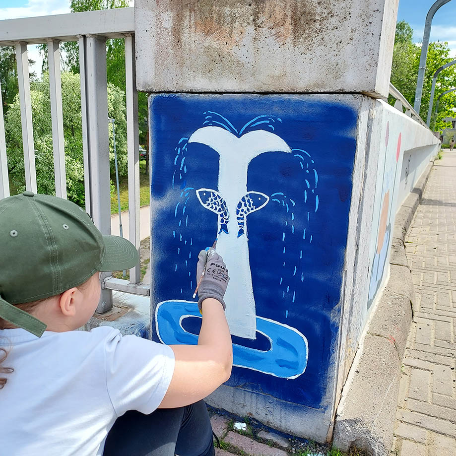 Kyykyssä oleva oppilas maalaa siveltimellä sinistä valaan pyrstöä esittävää maalausta betoniseen tolppaan