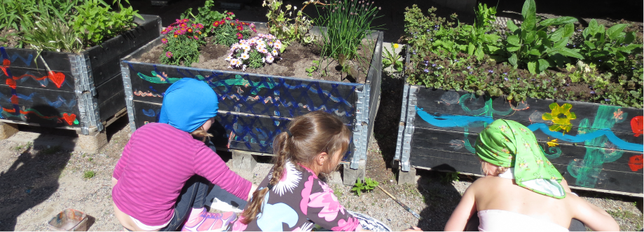 Kolme lasta selkäpuolelta kuvattuna, istuvat maassa ja maalaavat väreillä puisia laatikoita joissa kasvaa erilaisia kasveja ja kukkia.
