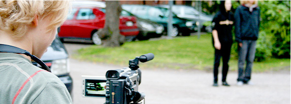 Selkäpuolelta kuvattu nuori, joka kuvaa videokameralla kahta muuta nuorta jotka seisovat kauempana. Taustalla puita, autoja ja tiilirakennus.