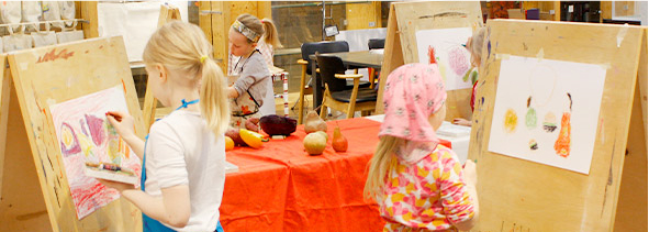 Kolme lasta piirtää hedelmä- ja vihannesasetelmaa värikynillä papereille, jotka ovat maalaustelineillä. Keskellä pöytä jossa on oranssi pöytäliina ja malliasetelma .