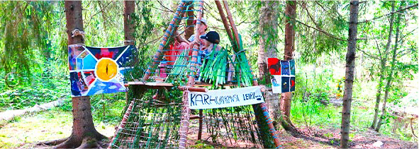 Kolmiomainen majarakennelma metsässä. Lapsia  istuu majassa. Puihin on kiinnitetty värikkäitä teoksia. Majaan on kiinnitetty kyltti jossa teksti: Karhunkynsi-leiri.