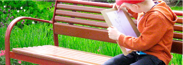 Oikeassa reunassa lapsi istuu puistonpenkillä ja piirtää sylissään olevalle paperille. Hänellä on oranssi huppari ja punainen lippis. Taustalla vihreitä kasveja.