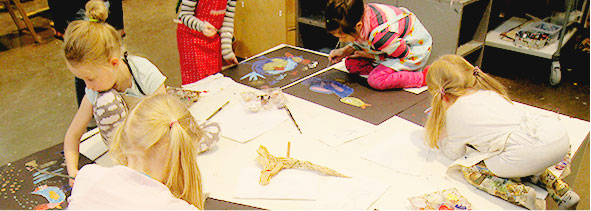 Neljä lasta istuu korokkeen päällä. Tekevät ruskeille papereille värikkäitä lintuaiheisia maalauksia.