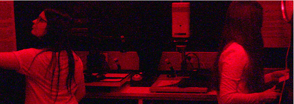 Pimeässä huoneessa otettu punasävyinen kuva, kaksi nuorta työskentelee pimiössä.