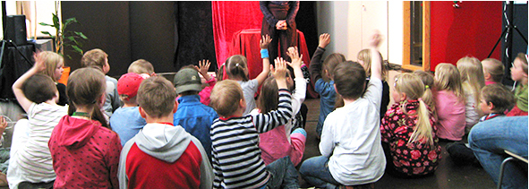 Selkäpuolelta kuvattuja lapsia jotka istuvat maassa ja katselevat edessä oleva esiintyjää. Osa lapsista viittaa kädellään.