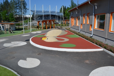 Rusetti-teos Kenttäkadun päiväkodissa. Harmaanvärisen päiväkotirakennuksen pihalla maassa on joutsenenmuotoinen veistos, minkä päällä voi istua ja kävellä.