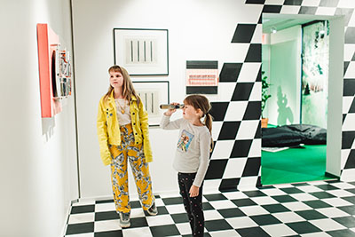 Kaksi lasta seisovat värikkäässä näyttelytilassa, Taidelabrassa. Toisella on kädessään kaukoputkimainen esine.