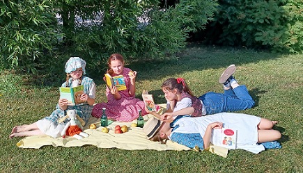 Kuvassa on nurmikolla viltillä neljä tyttöä, jotka ovat kukin pukeutuneet lukemansa tyttökirjaklassikon päähenkilöksi.