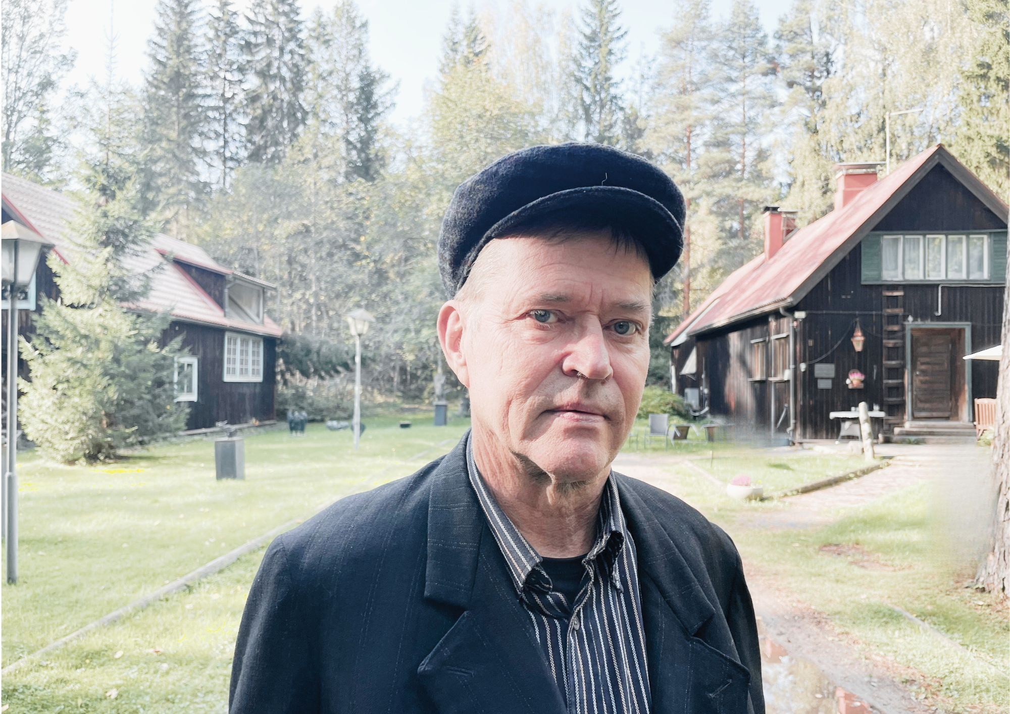 Valokuvassa mies katsoo kameraan vanhanaikaisessa lippalakissa, taustalla kaksi puutaloa.