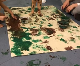 Kuvassa suuri sormiväreillä maalattu työ, yksi lapsi seisoo paljain jaloin työn keskellä lattialla ja maalaa jalkapohjillaan viidakon maisemaa