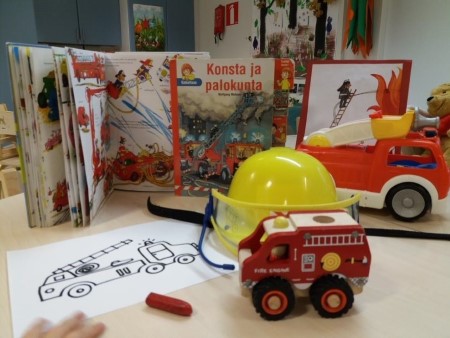 kuvassa on paloauto-aiheisia kirjoja pöydällä, leikkipaloauto, paloautovärityskuva ja keltainen palomiehen kypärä